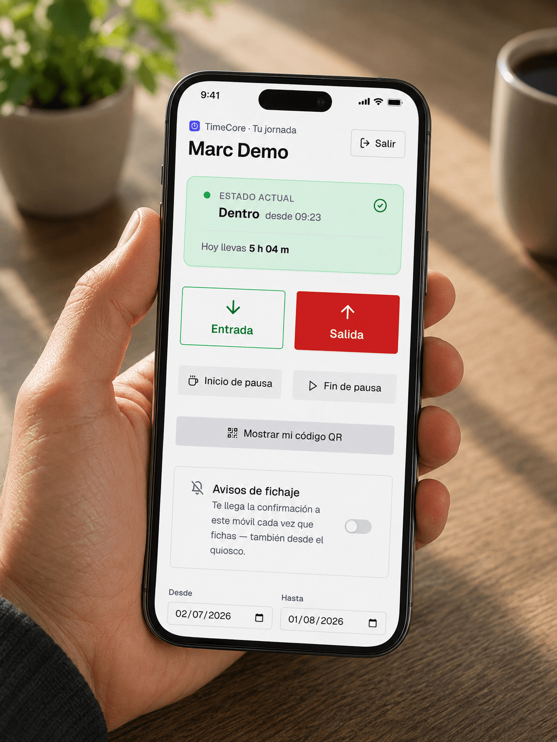 Una mano sostiene un móvil con el portal del trabajador de TimeCore: el nombre de la persona, la tarjeta «Estado actual — Dentro desde 09:23, hoy llevas 5 h 04 m» y los botones de Entrada, Salida, Inicio de pausa, Fin de pausa y «Mostrar mi código QR».