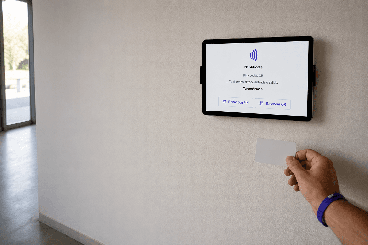 Una tablet montada en la pared junto a la entrada muestra la pantalla de espera del fichador de TimeCore —«Identifícate», «Fichar con PIN» y «Escanear QR»— mientras una mano le acerca una tarjeta lisa.